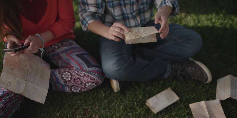 A young couple is outside reading letters and sitting on the grass