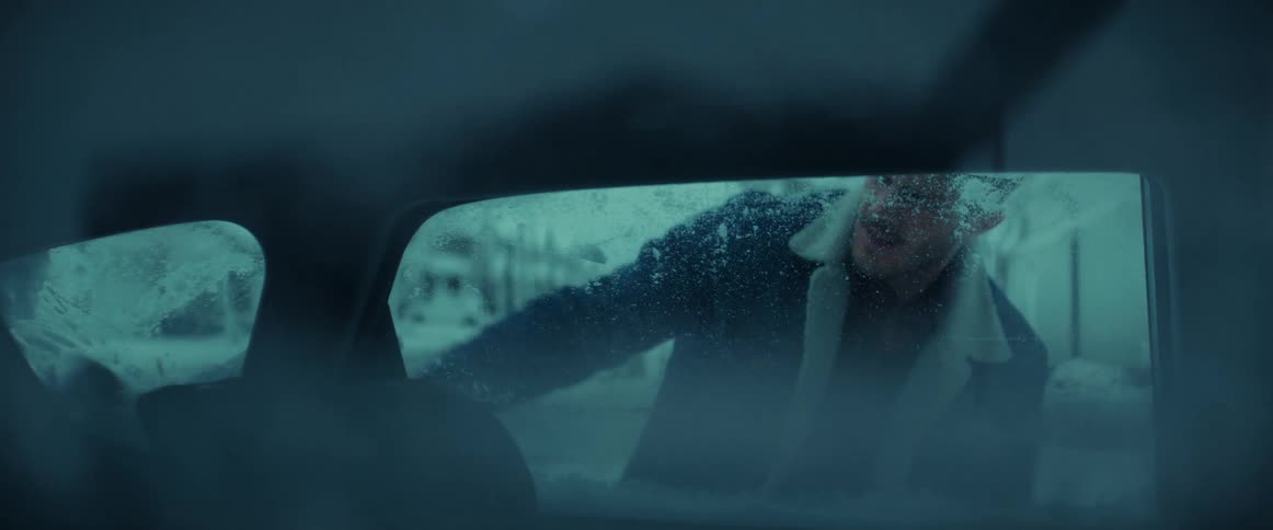 A man is standing outside holding a snowbrush and cleaning the snow from his car in his driveway