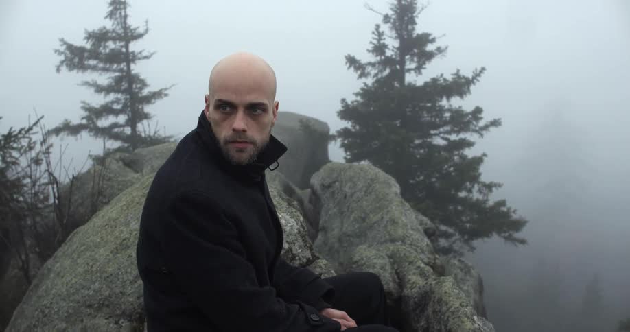 A man wearing a jacket sits atop a cliff overlooking a foggy forest