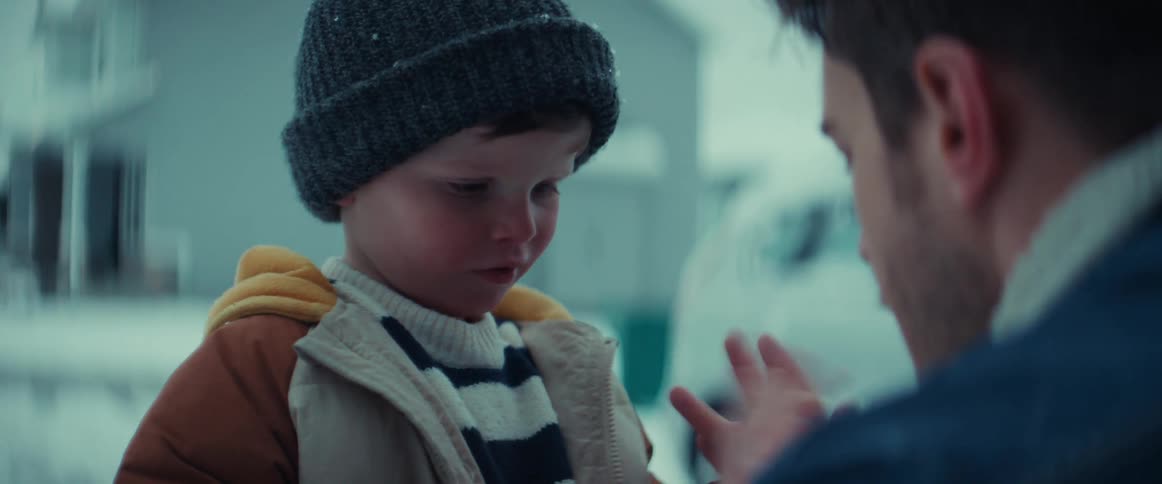 Father adjusts sons coat and blows on the childs hand to warm it as they stand near car on snowy day