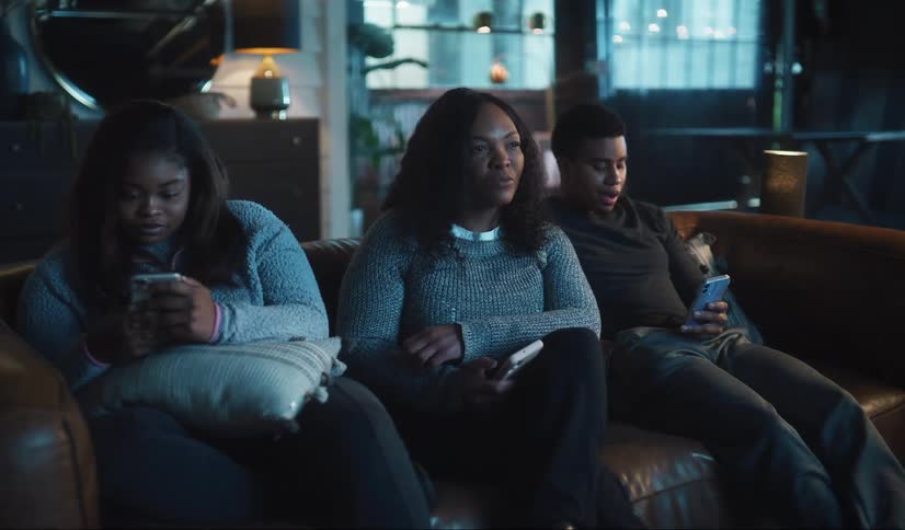 A mother is sitting on the couch with her teenage kids as they look on their phones