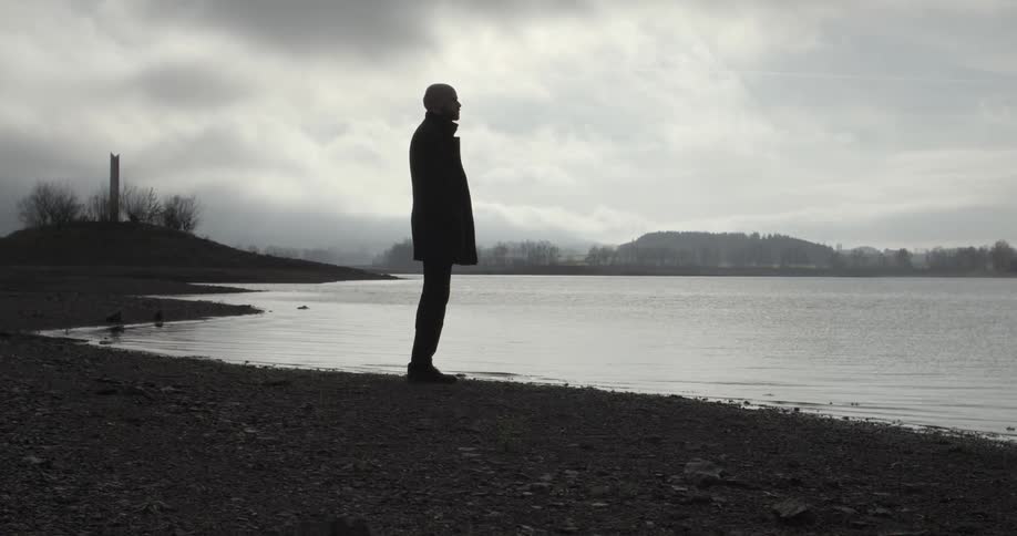 A man standing on a rocky shoreline under a cloudy, evening sky