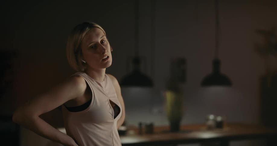 Blonde woman with tattoo sports bra shirt and earphones rides exercise bike near the kitchen