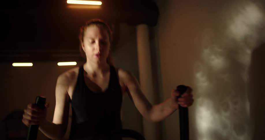 Redheaded woman smiles while exercising on elliptical in dimly lit room