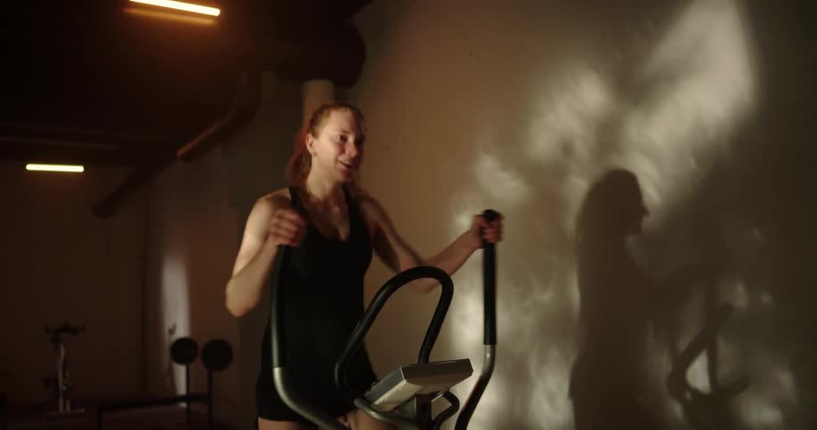 Determined woman with ponytail black top and shorts uses cross trainer at gym