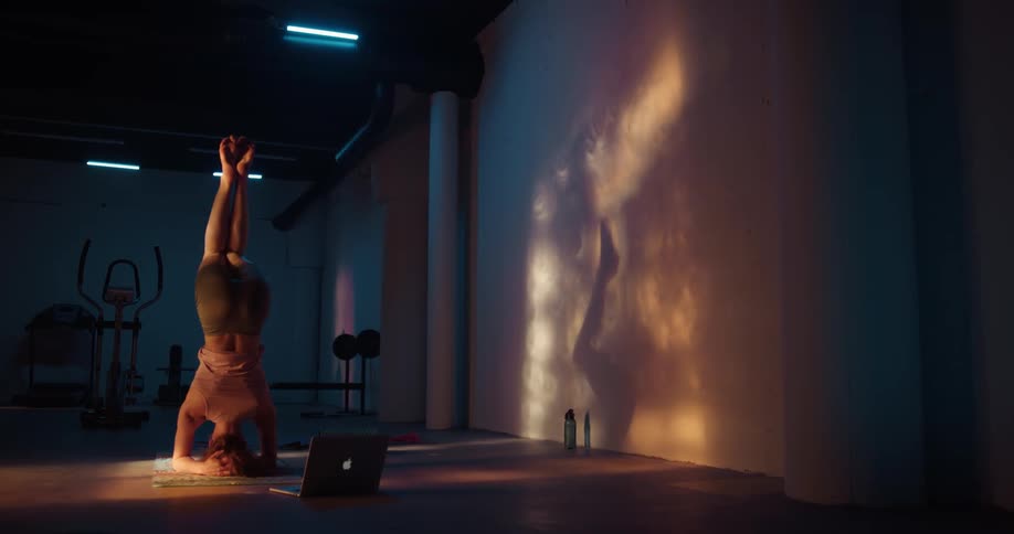 Redhaired woman balances on her head while performing yoga in dim home gym