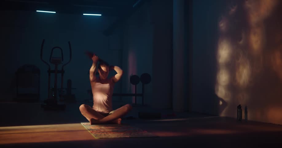 Woman sitting cross legged on yoga mat in dim room puts her hair into a ponytail before stretching her neck
