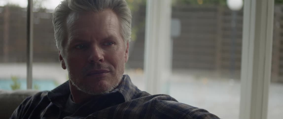 A man sitting on a sofa indoors looks at something out of frame with a serious expression