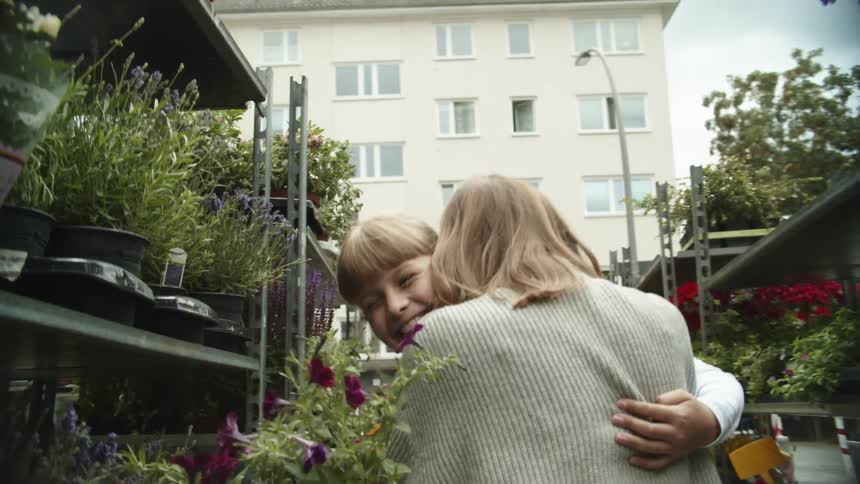 A little girl holding a pot with purple flowers runs to her mother down an aisle in a plant nursery at daytime