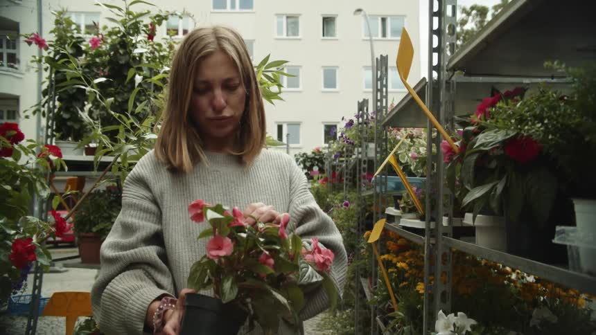 A woman checks out pink flowers in a pot while standing outside in a nursery next to other potted plants at daytime