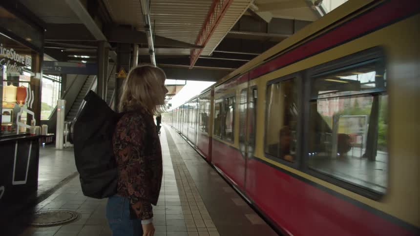 A woman stands at a train station and adjusts her backpack as a train pulls up