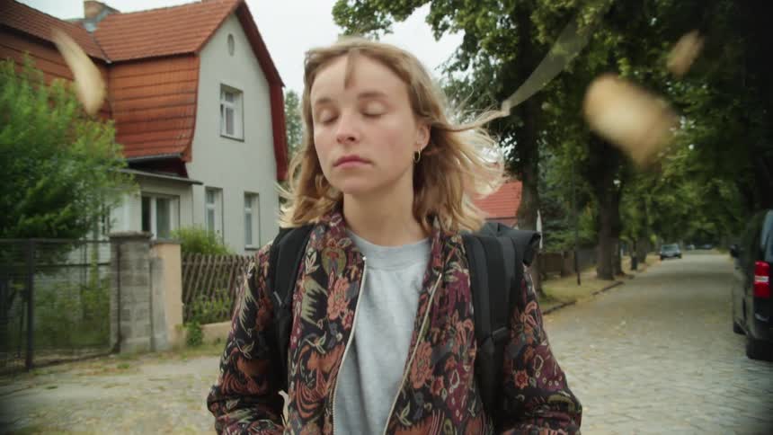 A woman with a jacket and backpack walking in a neighborhood with houses and trees the sky and clouds in the background