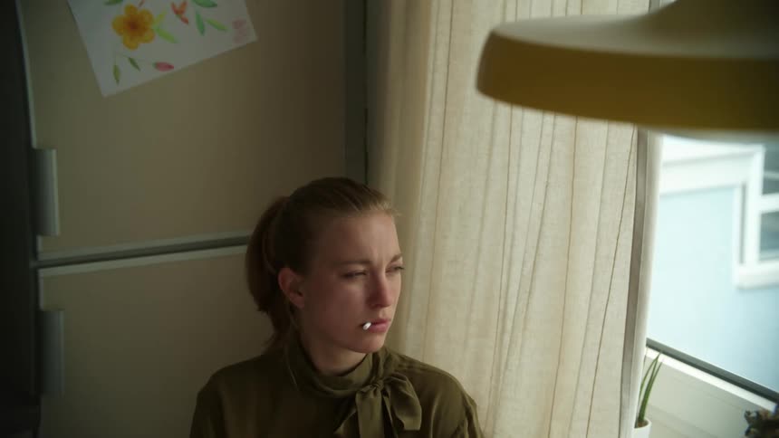 Young woman sitting in the kitchen at home near a window puts a cigarette on her mouth