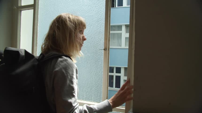 Young woman wearing a backpack opens a small window pane in a room