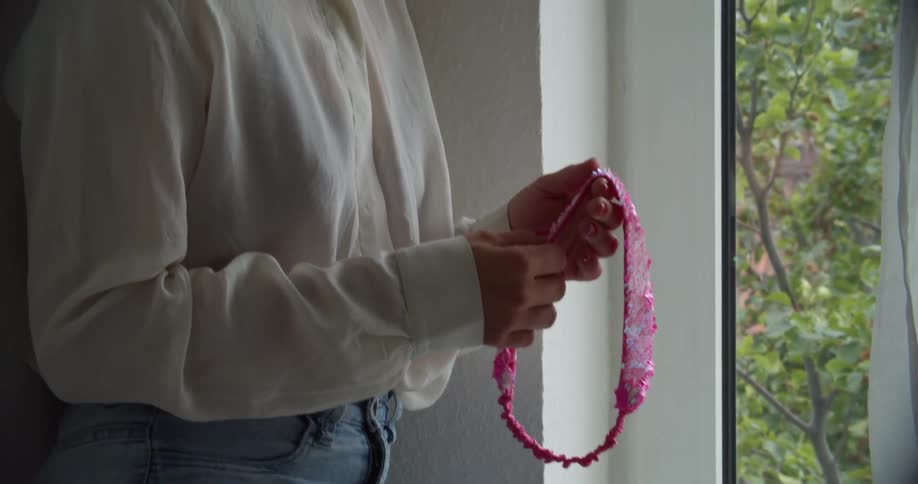 Woman playing with a pink accessory looking out the window of a room pensively