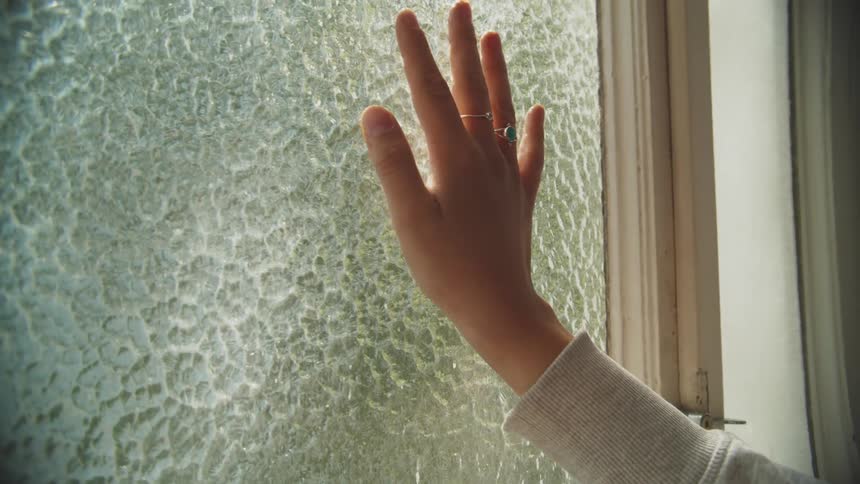 A female hand with rings on thefingers resting over a window pane inside a room