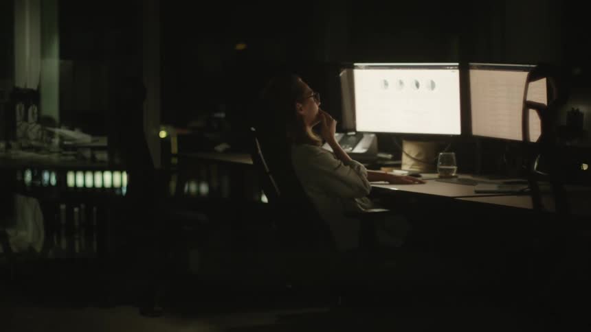 Tired woman with brown hair and light blue shirt sits back in front of computer screen in the office