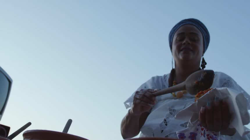 A woman is standing outside near a table with pots and she takes a wooden spoon from a bowl and adds a portion to the typical Bahia food in her hand Acaraje and looks at a monitor on a table