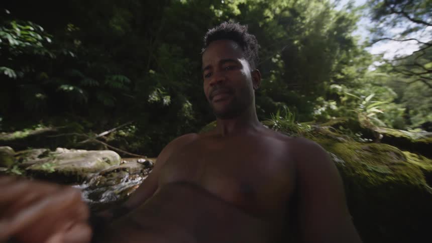 Topless man with orange shorts reads by the stream in the jungle