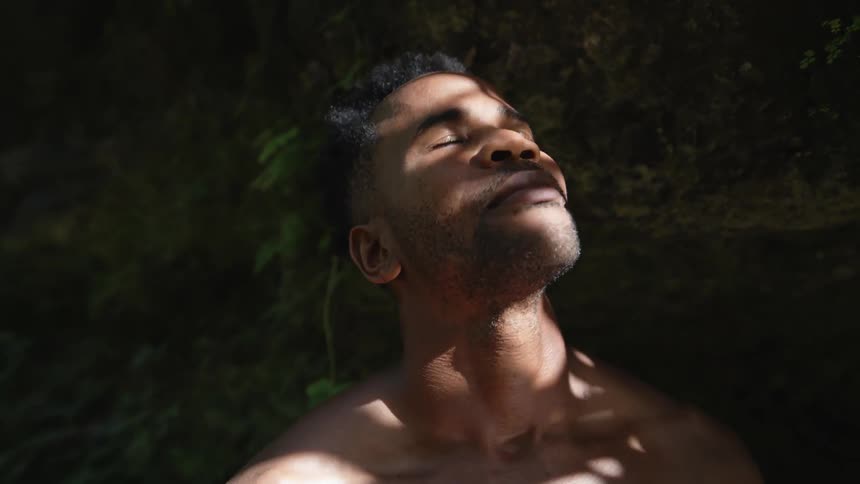 A man stands in front of a large rock with his eyes closed and lifts his head up towards the sky as the sun shines down on him