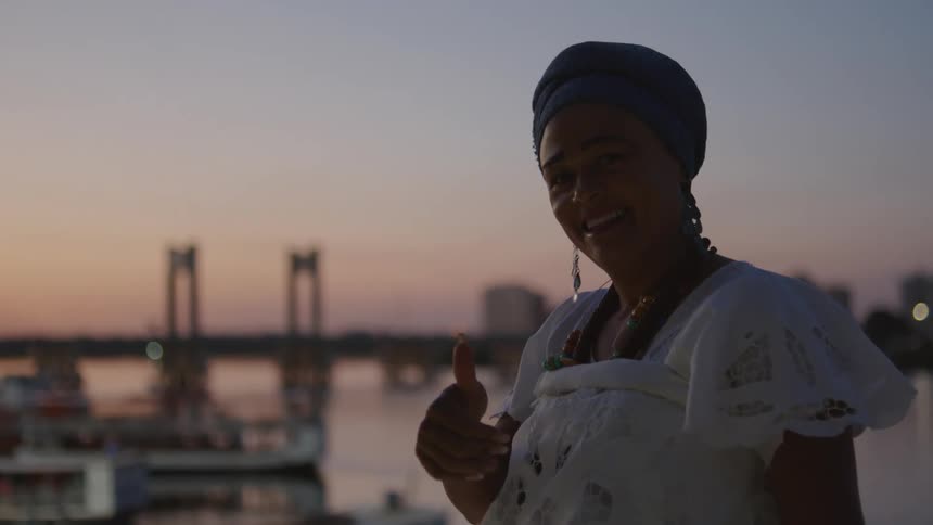 A woman looks at the view of a bridge then turns and smiles with her thumb up
