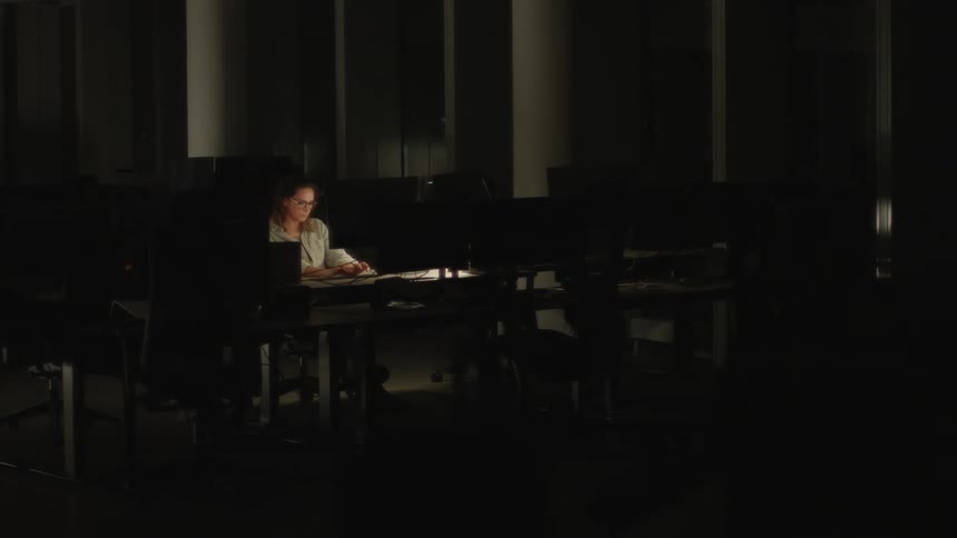 A woman is sitting in a mostly dark office typing on a computer
