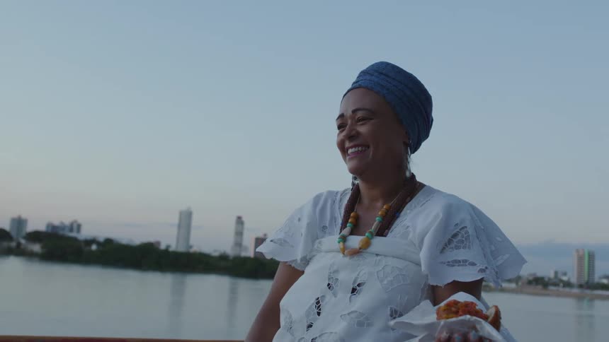 A woman in a white dress is standing near water laughing as she holds typical Bahia food Acaraje in a napkin in her hand