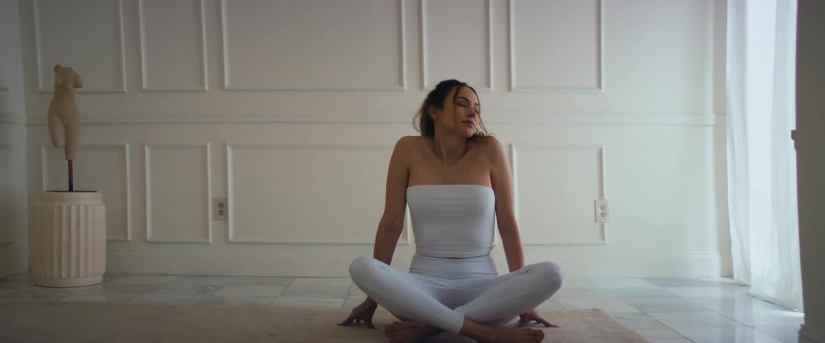 A thin woman walks into a room and sits with her legs crossed while stretching
