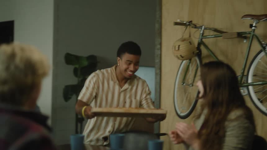 A man trips as he brings a pizza on a wooden board over to a group of 4 friends sitting at a table in front of a bicycle hanging on a wall
