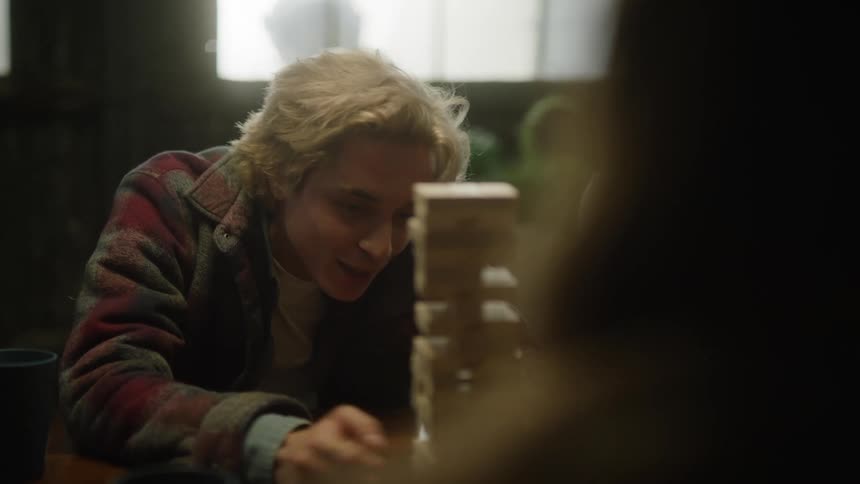 Young man wearing coat laughs and talks while playing stacked wooden blocks game