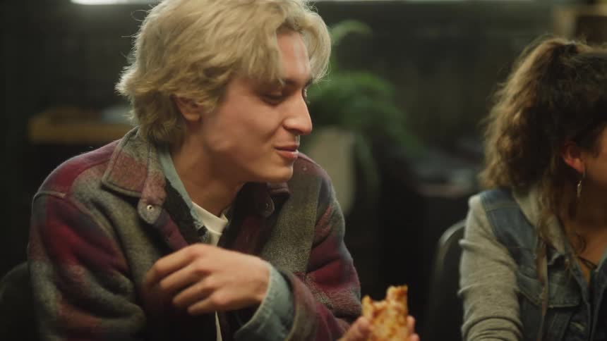 Blonde man and woman with brown wavy hair sit at desk eating pizza in room