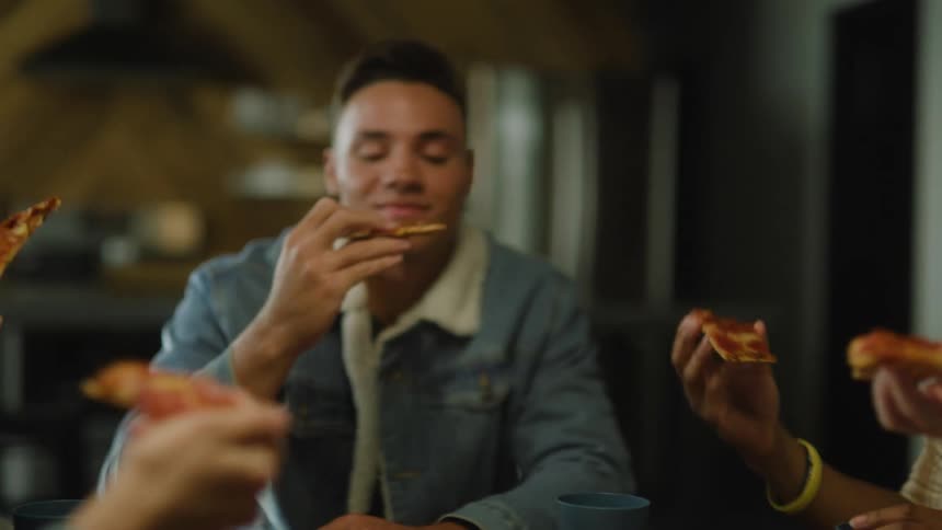 A small group of people are sitting at a table eating pizza in a restaurant and they raise their pizza slices in a toast