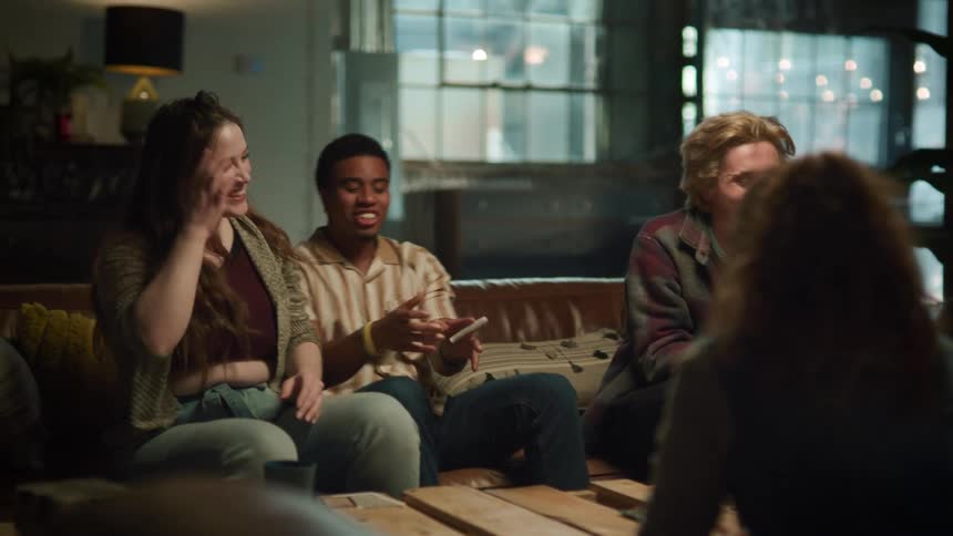 A group of friends sitting on couches talks to each other and laugh while playing cards in the living room