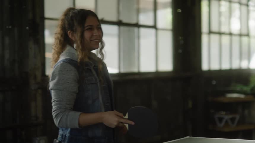A girl point her pingpong racket at someone else and laughing before hitting the pingpong ball