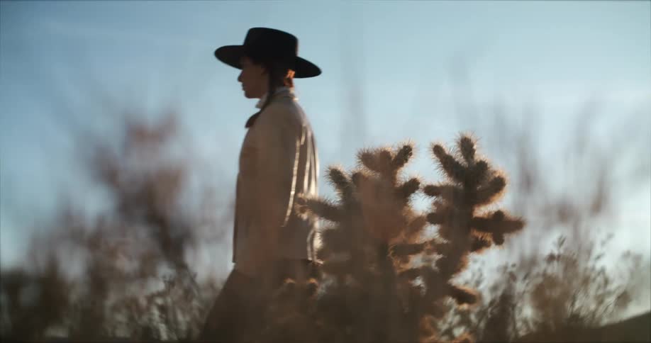 The sun shines brightly as a woman wearing a black hat walks among plants and trees under a clear sky