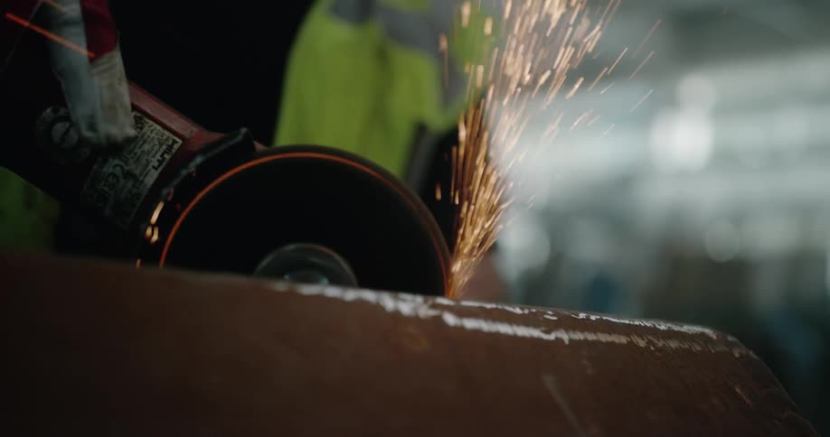 Worker wearing protective gear moves powered disk grinder over metal sending out sparks