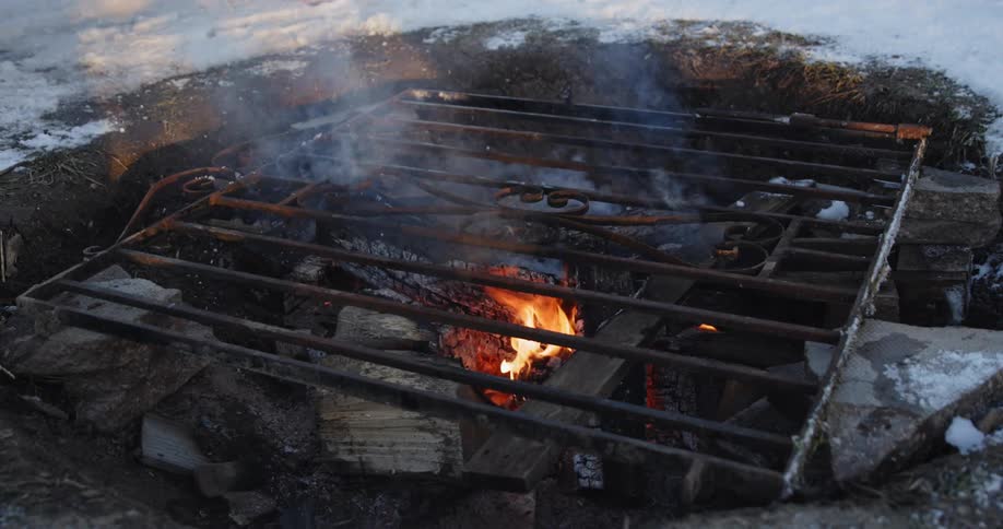 Charred wood is burning in a homemade grill outside in a firepit surrounded by snow