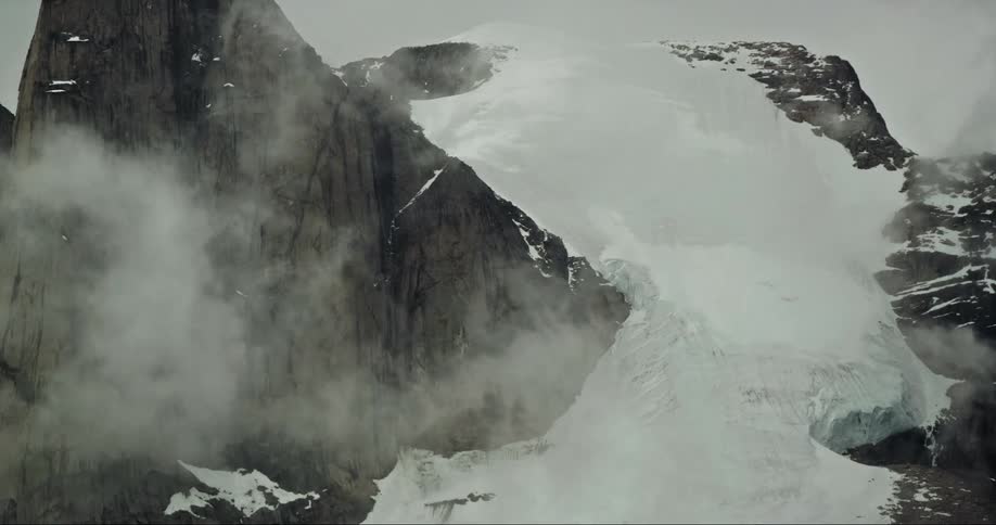 Clouds swirl above a giant glacier clinging to a granite alpine peak on Baffin Island