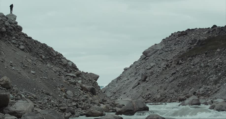 A person stands silhouetted on a steep and exposed rocky ravine by a raging river