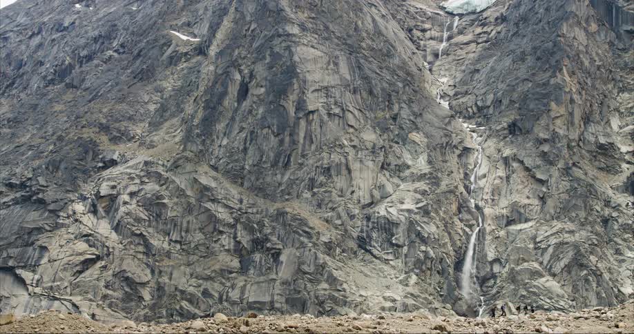 Hikers walking by a waterfall and a towering granite cliff face in the background