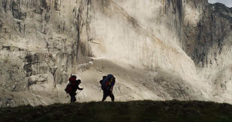 Two silhouetted people trekking under the towering cliff face of Mount Thor in Auyuittuq National Park on Baffin Island