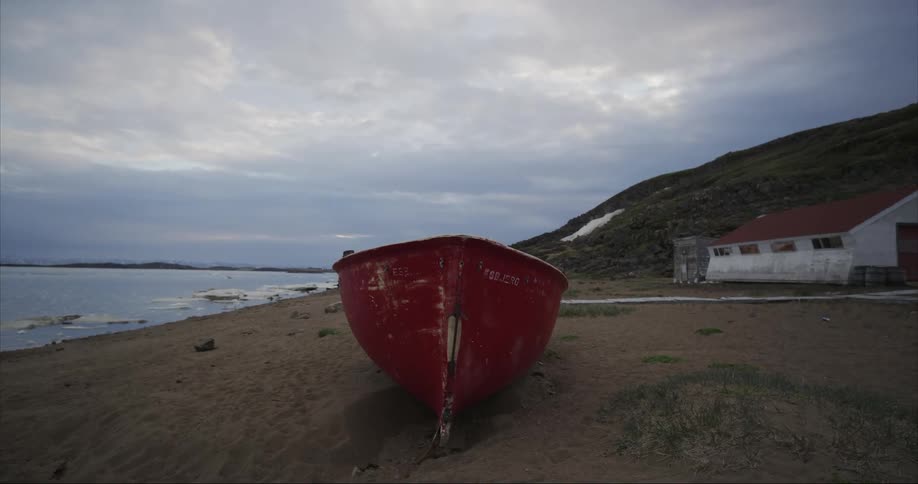 View of a beat up beached boat with red hull under cloudy skies white house with red roof on the coast of the arctic ocean on Baffin Island