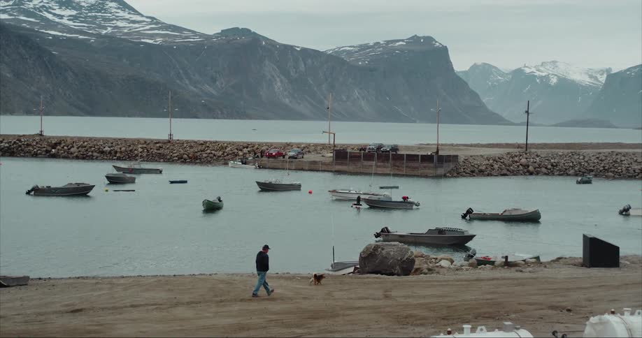 A man walking next to a sea harbor with boats with snowy mountains in the distance on Baffin Island