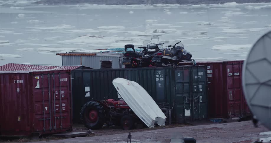 Shipping containers, boats, a tracor and snowmobiles while sea ice swirls in the background