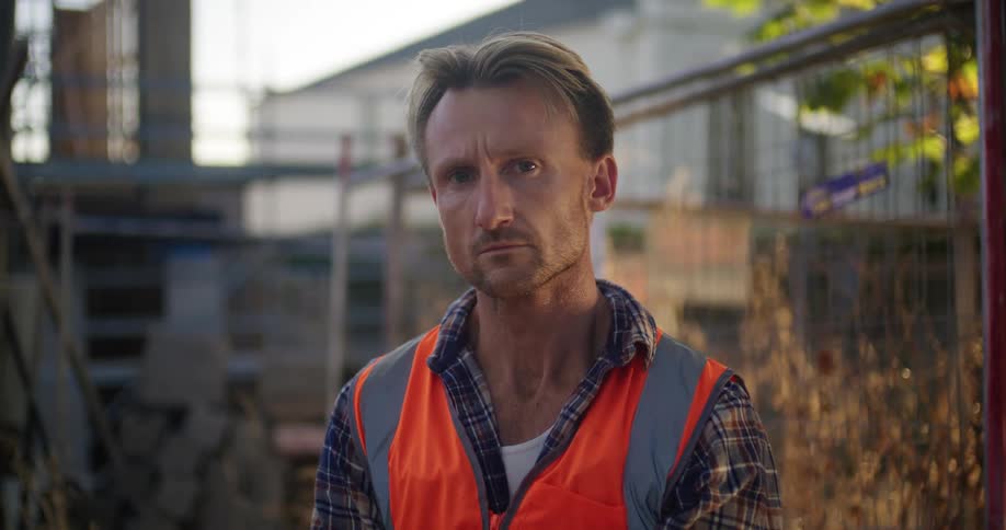 A man wearing an orange vest is standing in front of a bunch of wooden planks outside