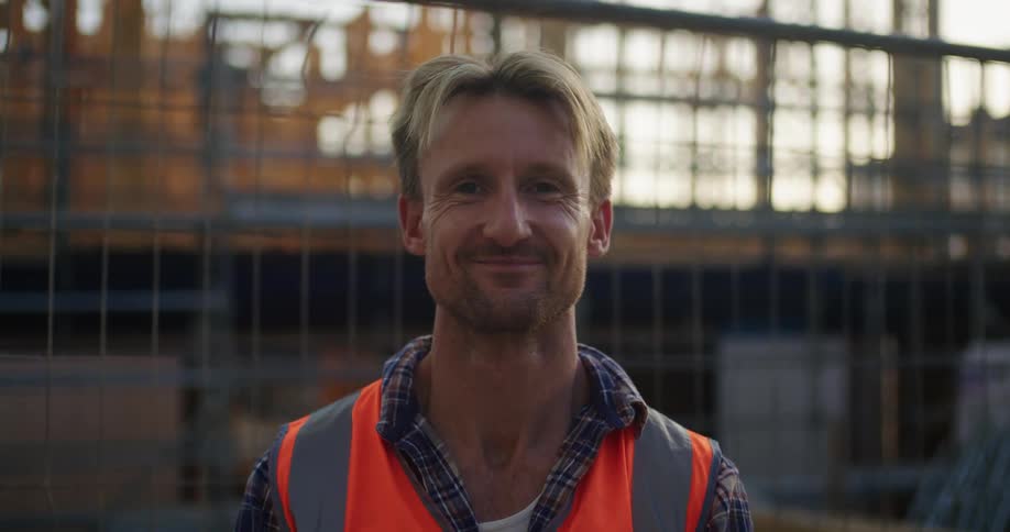 Blonde man wearing plaid shirt and safety vest smiles at camera at construction area