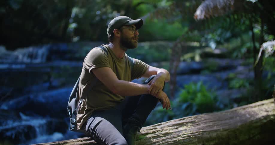 A man out hiking rests on a log by a waterfall and looks at his cell phone