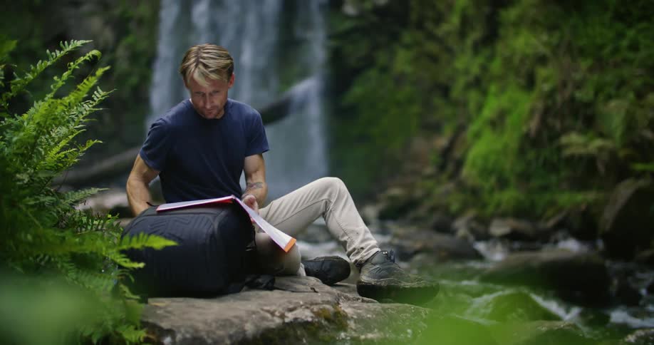 Man sitting on riverbank near waterfall flips through folder after using device to check area