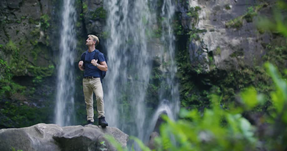 A man with a backpack on is standing in front of a waterfall over a cliff and standing on a rock
