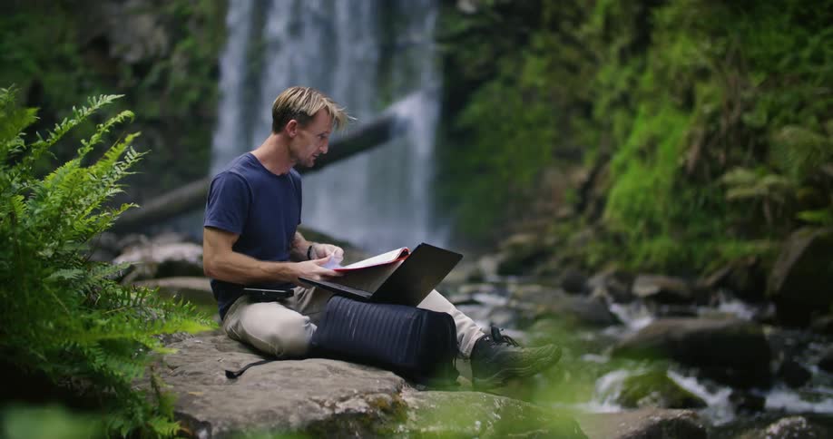 A man is typing on his laptop and flipping through papers by the waterfall